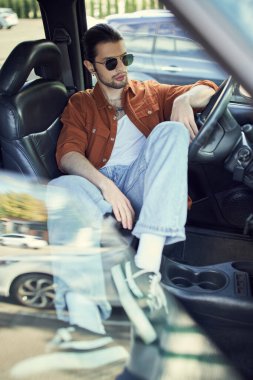 Rahat, güneş gözlüklü, kot pantolonlu, kahverengi gömlekli, arabasında takılan, moda anlayışı olan çekici bir adam.