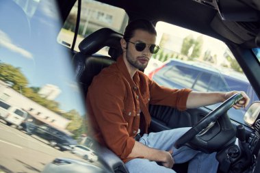 young good looking man with stylish sunglasses in vibrant attire posing in car and looking at camera clipart