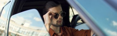 young stylish man with trendy accessories posing behind steering wheel, touching sunglasses, banner clipart