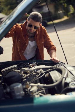 Aksesuarlı kahverengi gömlekli çekici genç adam arabasının motoruna dikkatle bakıyor.