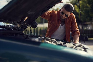 Arabasının motoruna bakan at kuyruklu ve modaya uygun aksesuarlı düşünceli erkek model.
