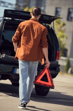 Uyarı üçgenini elinde tutarak arabasına doğru yürüyen şehir kıyafetli genç bir adamın arka görüntüsü