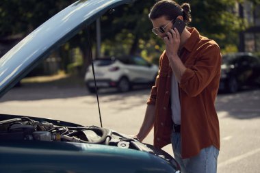 Kahverengi gömlekli yakışıklı seksi adam sigortacısıyla konuşuyor ve arabasının motoruna bakıyor.