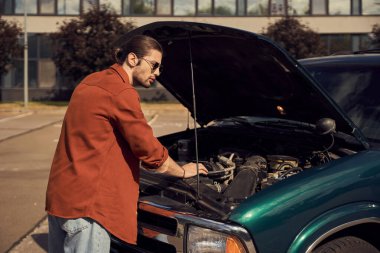 handsome bearded man with ponytail and sunglasses standing next to his car with opened engine hood clipart