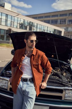 Yakışıklı, seksi, şık görünümlü, arabanın yanında duran ve başka yöne bakan erkek modelin dikey çekimi.