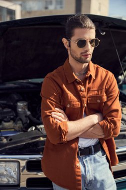 Yakışıklı göz alıcı erkek model şık giyinmiş, kolları göğsünde arabasının yanında poz veriyor.