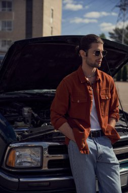 vertical shot of good looking young man in brown shirt with sunglasses posing near his broken car clipart