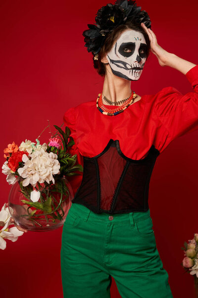 woman in skeleton makeup and festive attire holding vase with colorful flowers on red, Day of Dead