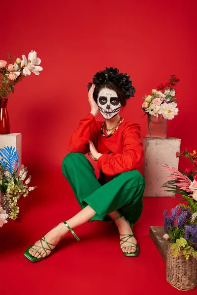 woman in skeleton makeup and black wreath sitting near dia de los muertos altar with flowers on red