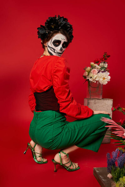 женщина в dia de los muertos макияж глядя на камеру возле традиционного алтаря с цветами на красном