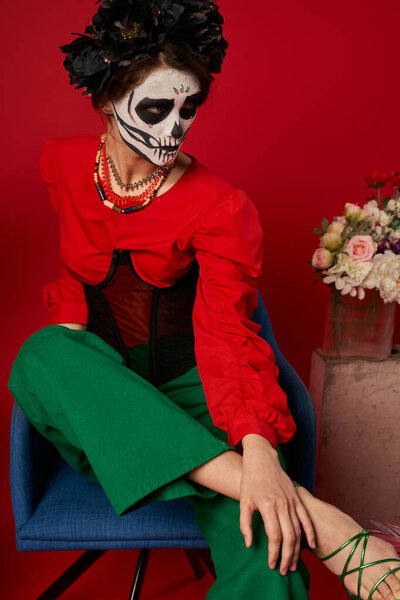 woman in catrina makeup sitting in armchair near flowers on red, dia de los muertos tradition