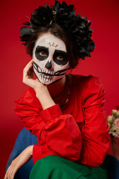 portrait of woman in skull makeup and black wreath looking at camera on red, Day of Dead tradition