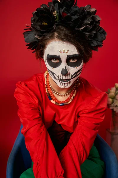 woman in scary sugar skull makeup and colorful beads looking at camera in armchair on red