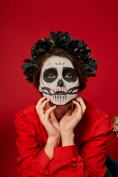 spooky woman in skull makeup and black wreath holding hands near face on red, dia de los muertos