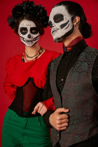 woman in skull makeup and black wreath looking at camera near man on red, dia de los muertos couple