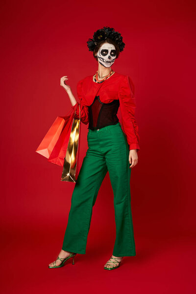 full length of woman with dia de los muertos makeup and shopping bags on red, seasonal sale