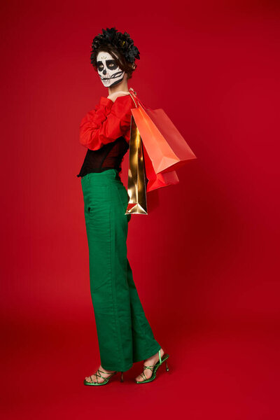 full length of woman in Day of Dead makeup and festive attire posing with shopping bags on red