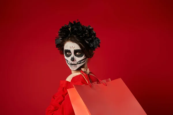 portrait of woman in sugar skull makeup and black wreath with shopping bags on red, seasonal sale