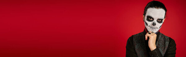 portrait of man in dia de los muertos sugar skull makeup holding hand near chin on red, banner