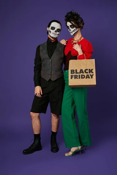 woman in sugar skull makeup and black wreath holding black friday shopping bag near man on blue