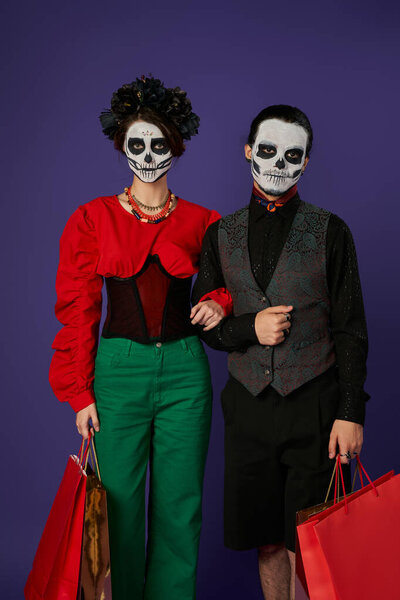 dia de los muertos couple in sugar skull makeup holding shopping bags and looking at camera on blue