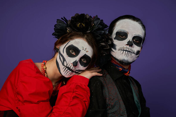 woman in sugar skull makeup and black wreath leaning on shoulder of eerie man on blue, Day of Dead