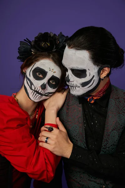 woman in skull makeup and black wreath leaning on shoulder of man and looking at camera on blue