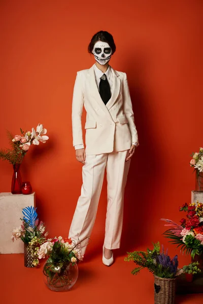 stock image woman in skull makeup and white suit near traditional dia de los muertos ofrenda with flowers on red