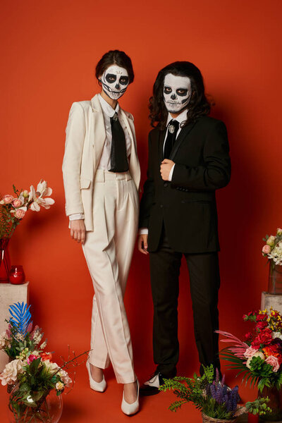 couple in sugar skull makeup and  elegant suits near dia de los muertos altar with flowers on red