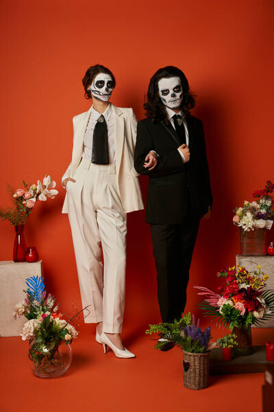 couple in catrina makeup and suits posing near dia de los muertos ofrenda with flowers on red