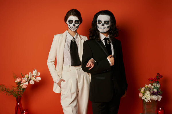 couple in catrina makeup and suits near festive dia de los muertos ofrenda with flowers on red