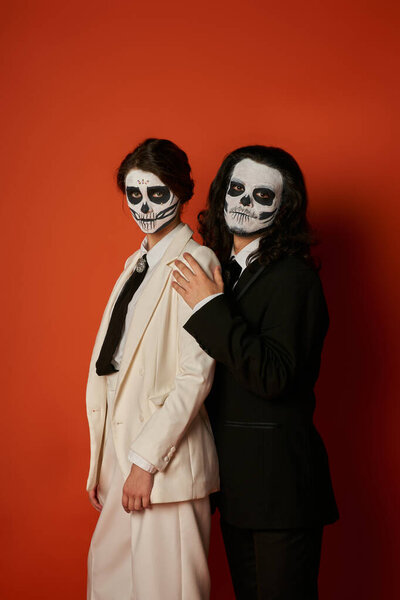 elegant couple in dia de los muertos makeup and elegant festive suits looking at camera on red