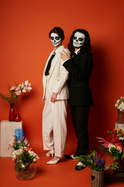couple in skeleton makeup and suits near festive dia de los muertos ofrenda with flowers on red
