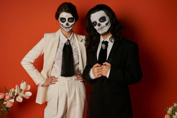 elegant couple in catrina calavera makeup looking at camera near floral decor on red, Day of Dead