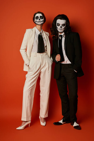 full length of stylish couple in skull makeup and elegant suits posing with hands in pockets on red