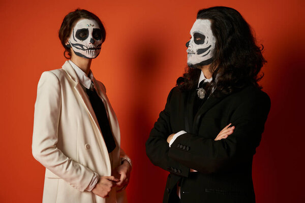 couple in dia de los muertos makeup, man with folded arms looking at woman in white suit on red