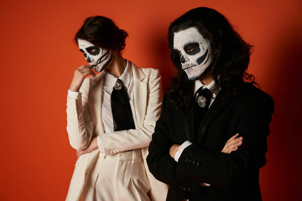 thoughtful couple in sugar skull makeup and elegant suits looking away on red, Day of Dead concept