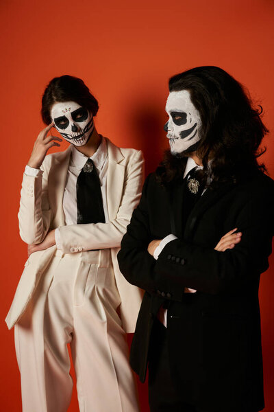 woman in catrina calavera makeup and white suit looking at spooky man with folded arms on red