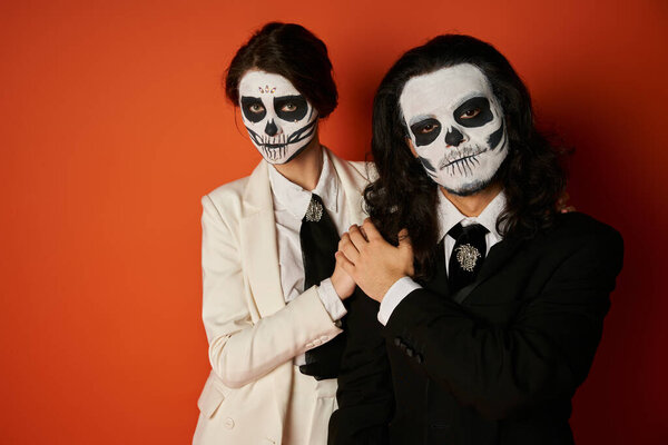 couple in dia de los muertos makeup and elegant suits holding hands and looking at camera on red