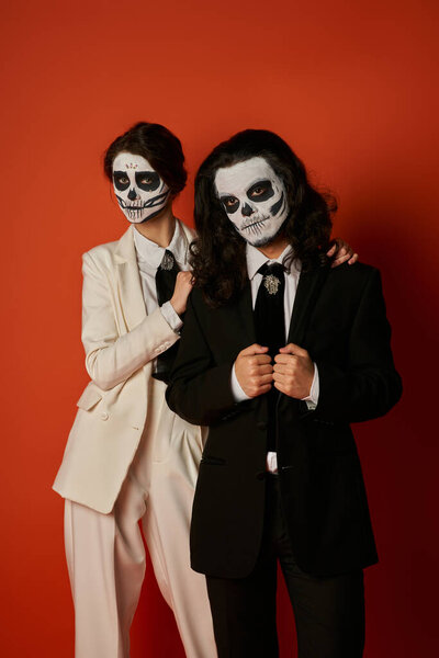 stylish couple in dia de los muertos sugar skull makeup and suits looking at camera on red backdrop