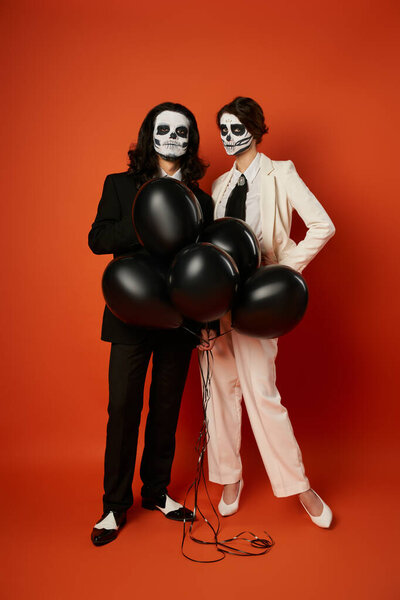 full length of couple in skull makeup and suits with black balloons on red, dia de los muertos party