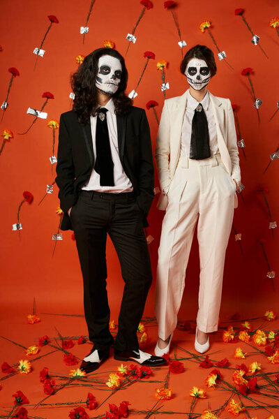 elegant couple in dia de los muertos makeup and suits posing on red backdrop with carnation flowers