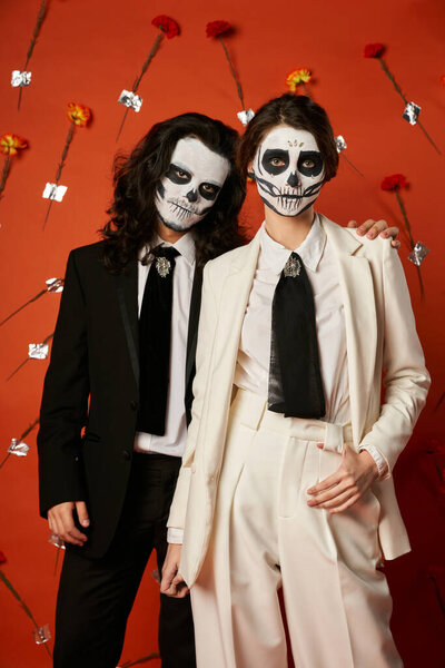 couple in dia de los muertos skull makeup and suits looking at camera on red backdrop with flowers