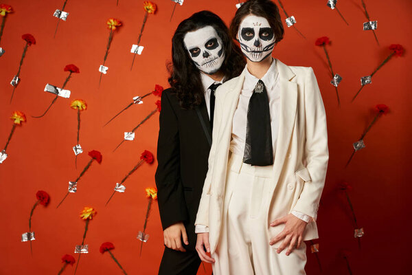 couple in dia de los muertos catrina makeup and suits looking at camera on red backdrop with flowers