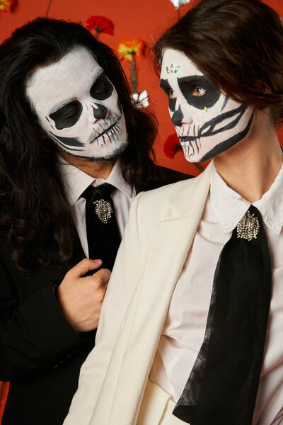 dia de los muertos couple, spooky man looking at woman in skull makeup on red backdrop with flowers