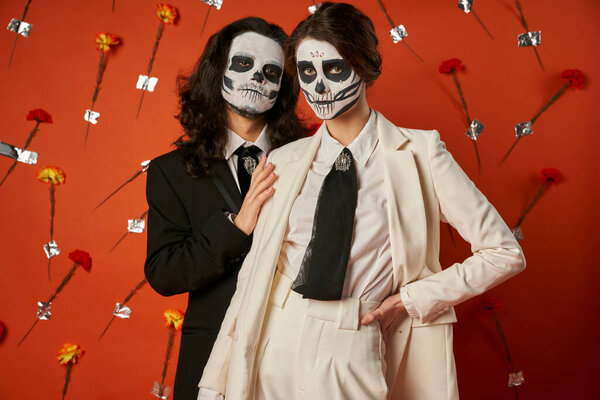 couple in scary sugar skull makeup and festive attire looking at camera on red backdrop with flowers