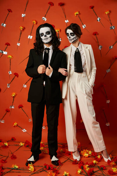 couple in catrina makeup and festive attire posing on red backdrop with carnations, full length