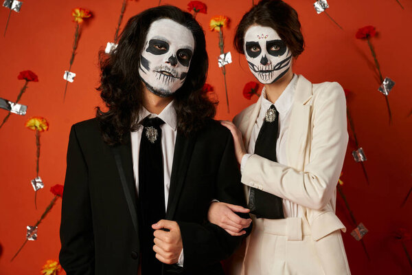 couple in eerie sugar skull makeup and festive attire looking at camera on red backdrop with flowers