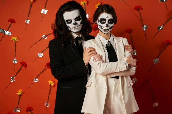 dia de los muertos couple, woman with folded arms near scary man on red backdrop with flowers