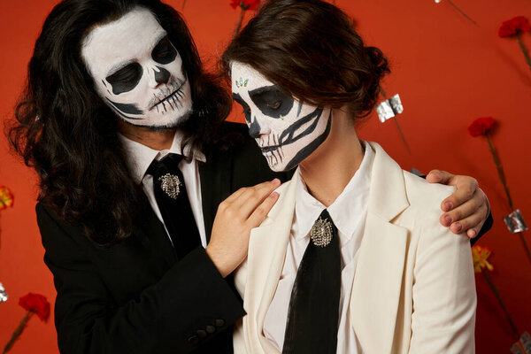 spooky man embracing shoulders of woman in suit, dia de los muertos couple on red floral backdrop